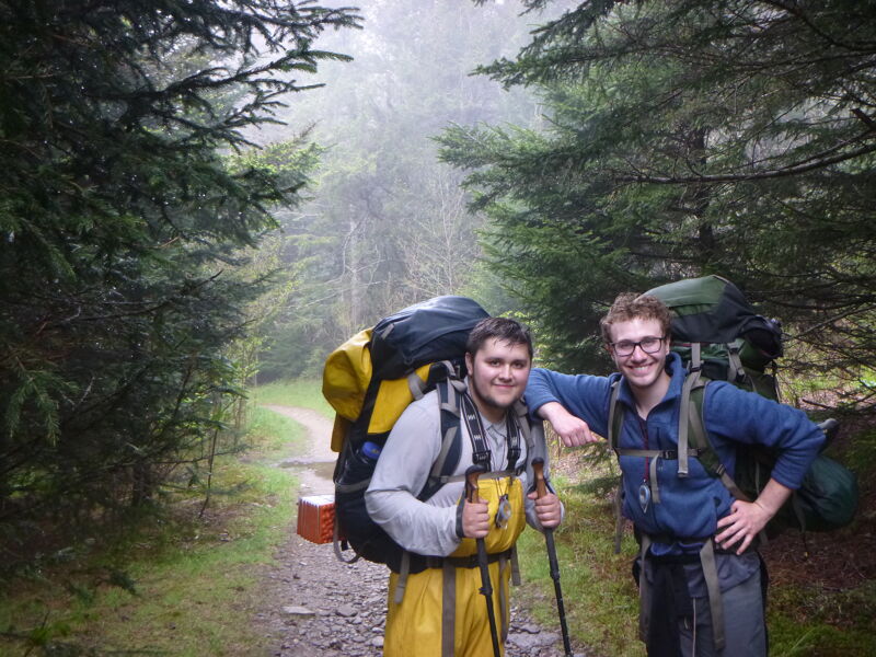 Two young men are standing on a trail in a forest, wearing large backpacks and hiking gear. The man on the left is wearing yellow rain pants and holding hiking poles. The man on the right is wearing glasses and has his hand on his hip. The trail is surrounded by trees and there is fog in the distance.